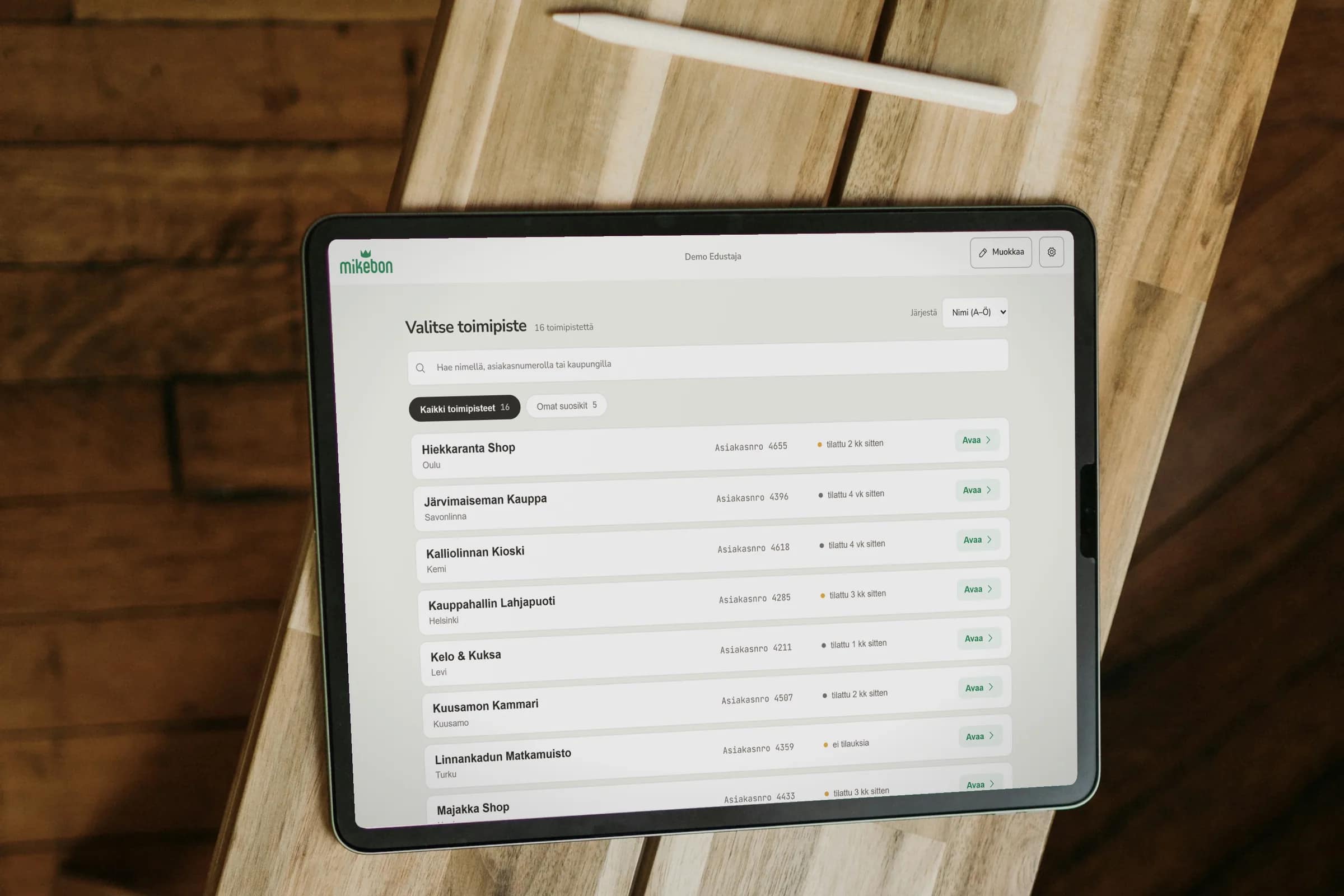 Seen from above, a tablet lying on a wooden bench next to a stylus, showing the sales reps’ store picker: a list of shops with customer numbers, how long ago each one ordered, and an Avaa (open) button on every row.