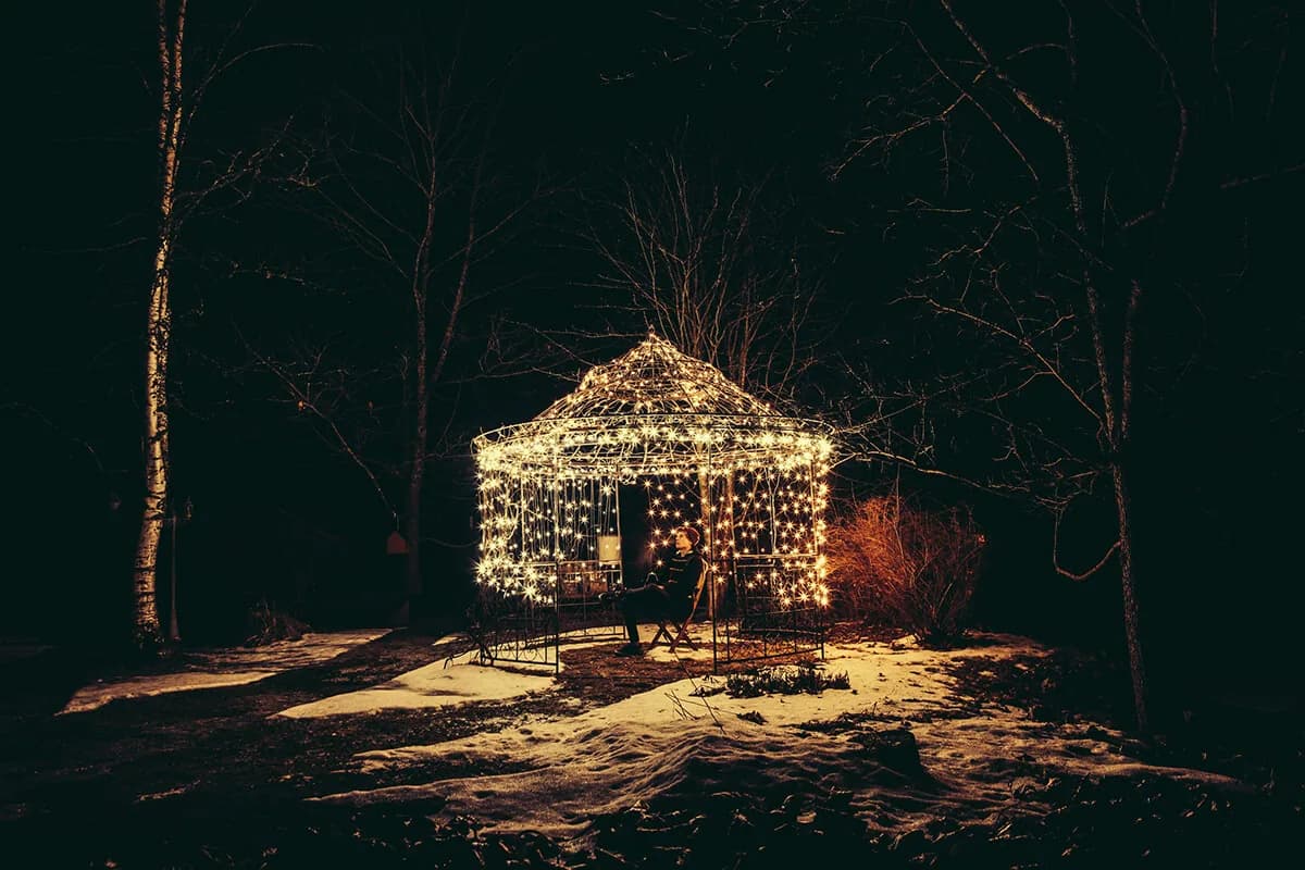 A gazebo covered in warm string lights in a dark winter garden, a person sitting inside, patches of snow on the ground.
