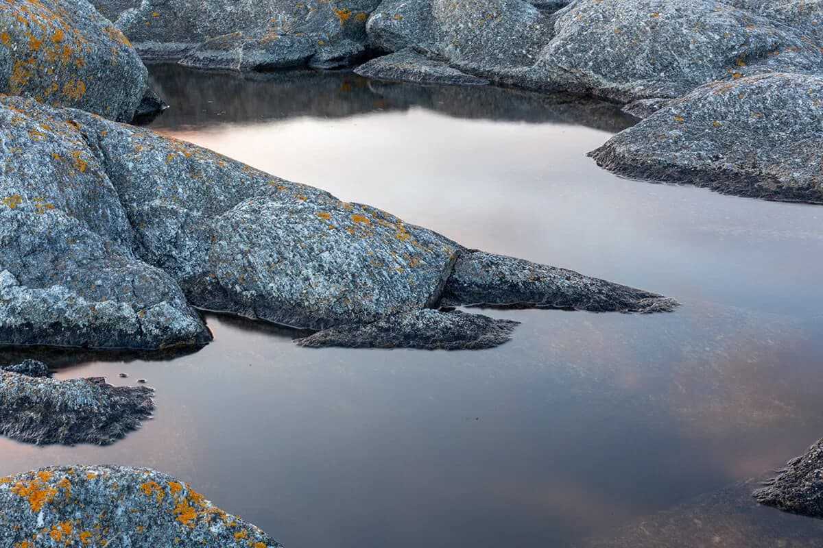 A still rock pool between grey granite slabs with orange lichen, reflecting a soft evening sky.