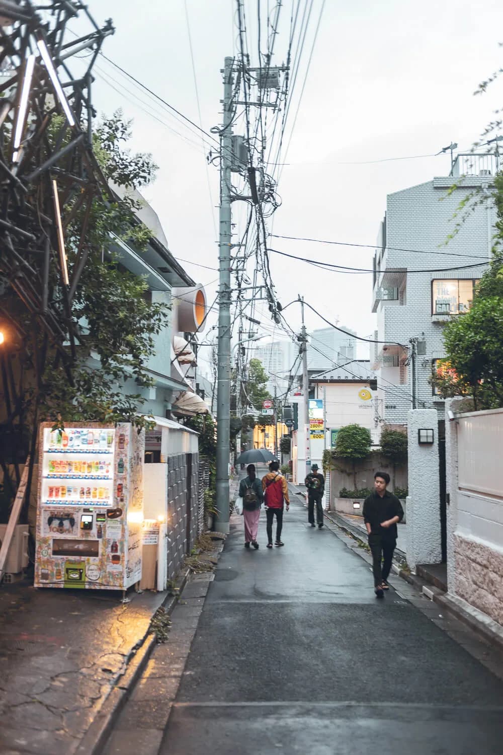 Narrow Tokyo side street on a grey evening: a lit vending machine on the left, tangled power lines overhead and a few people walking on wet asphalt.